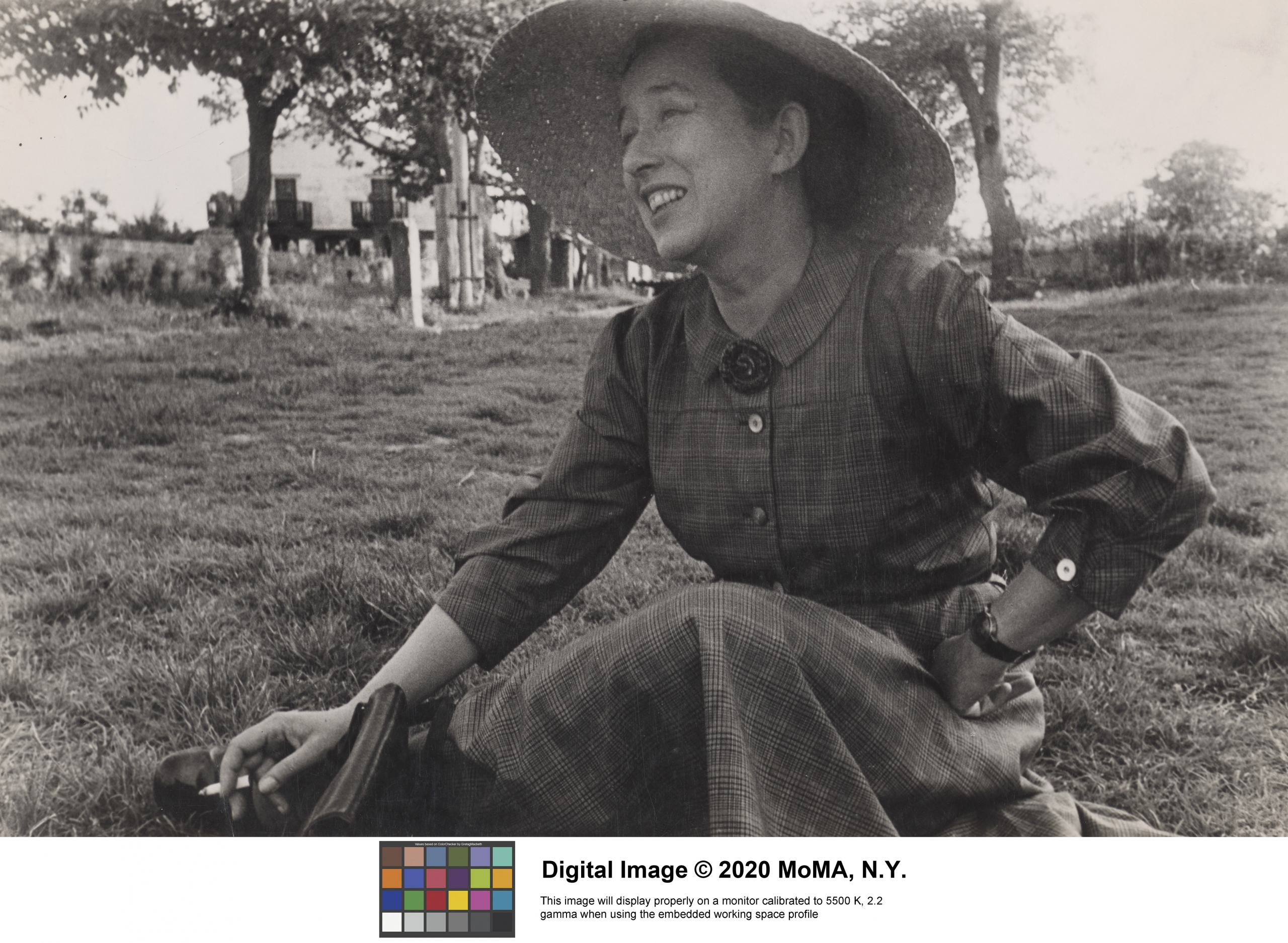 Archival photo of Margaret Scolari Barr sitting on the grass and smiling off camera. Barr is wearing a long-sleeve dress and large brim hat.