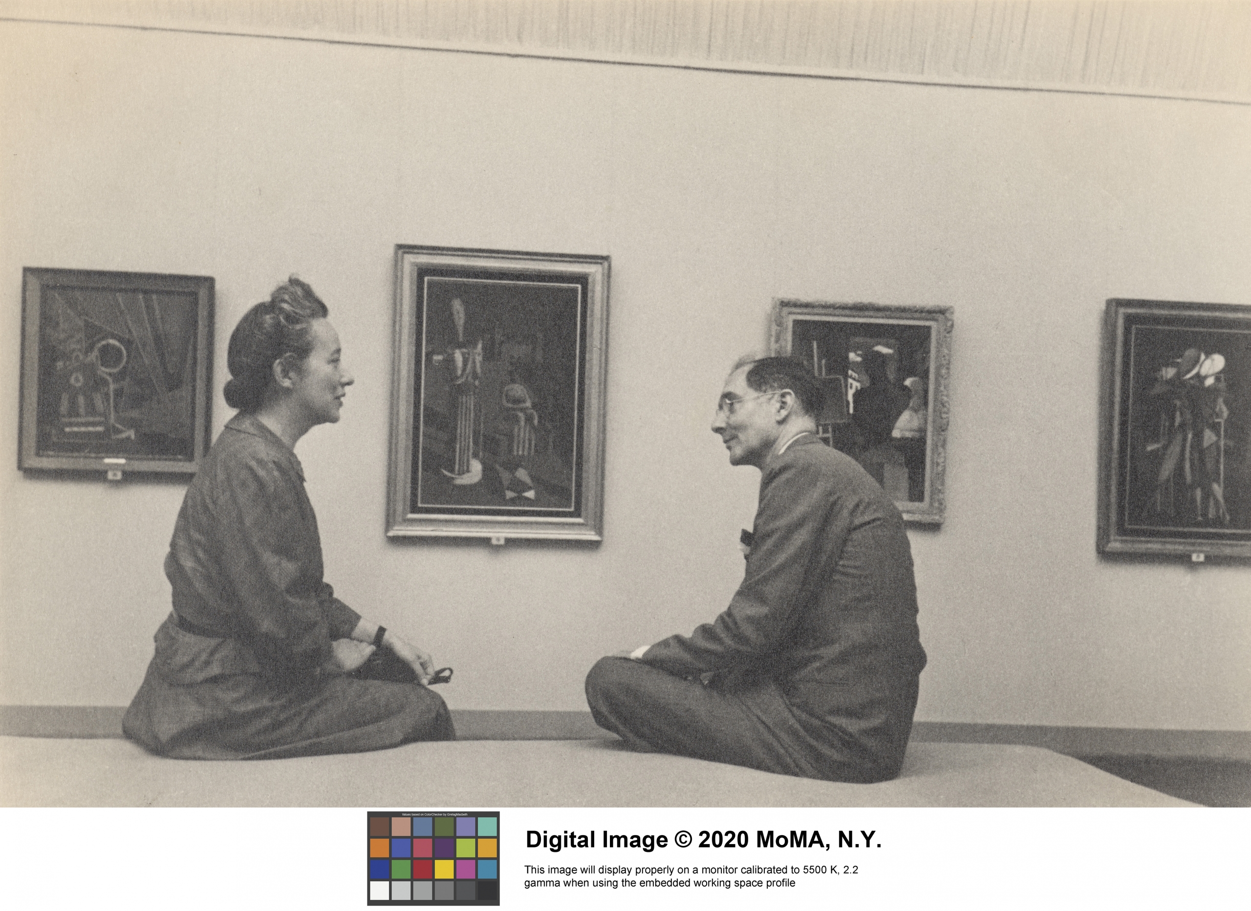 Archival photo of Margaret Scolari Barr and her husband Alfred H. Barr, Jr. The couple is sitting on a bench in a museum talking to each other. Behind them hangs four large paintings.