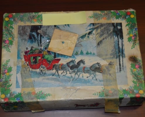 Photo of an old box, about the size of a shoe box, with letters inside. The box has an image of a horse-drawn carriage and holly leaves on it, with faded and peeling tape holding it in place.