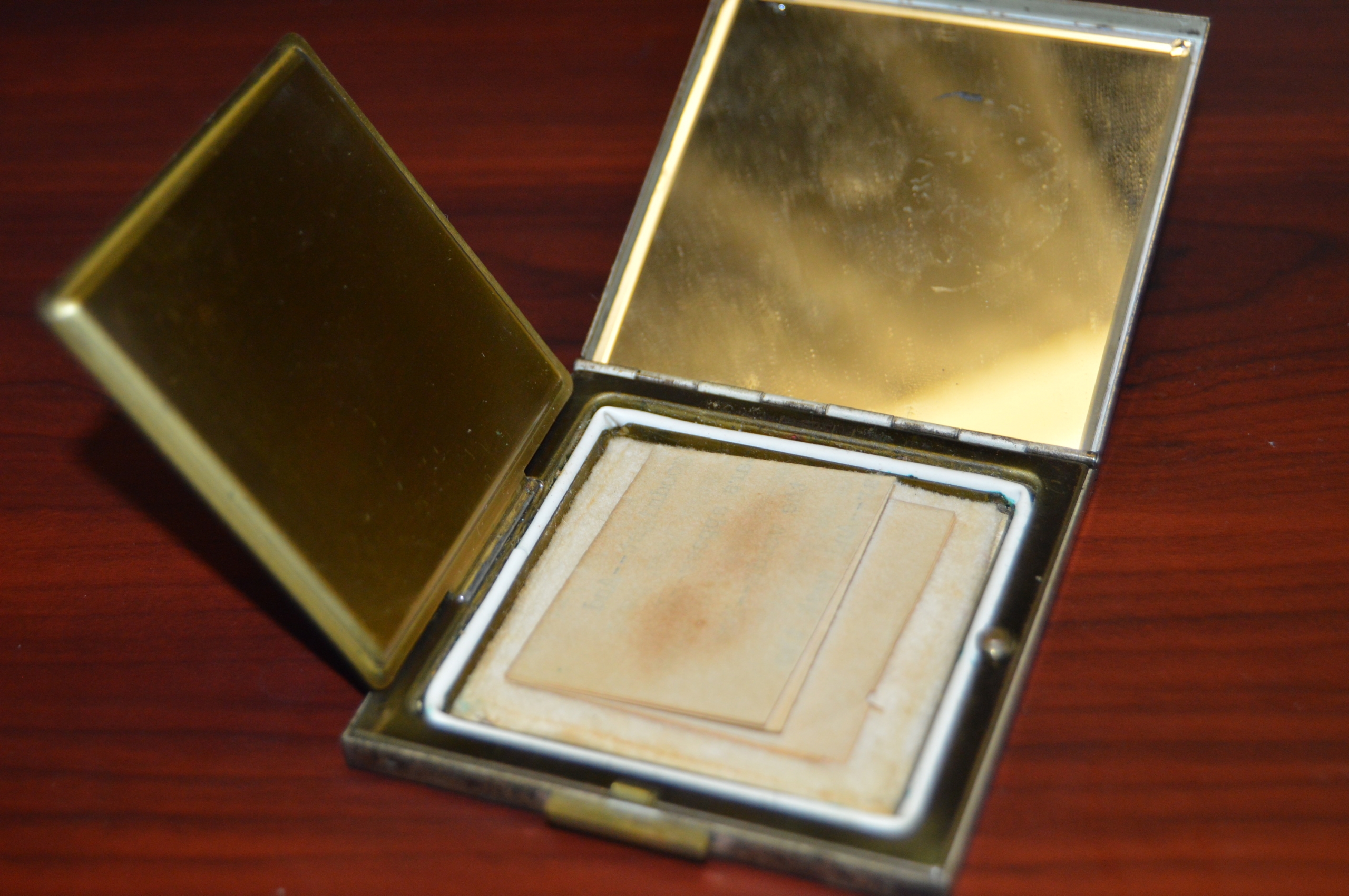 Photo of a small square compact that opens and unfolds from the top and left sides. The top side is a mirror. In the main compartment of the compact there appears to be small folded notes. 