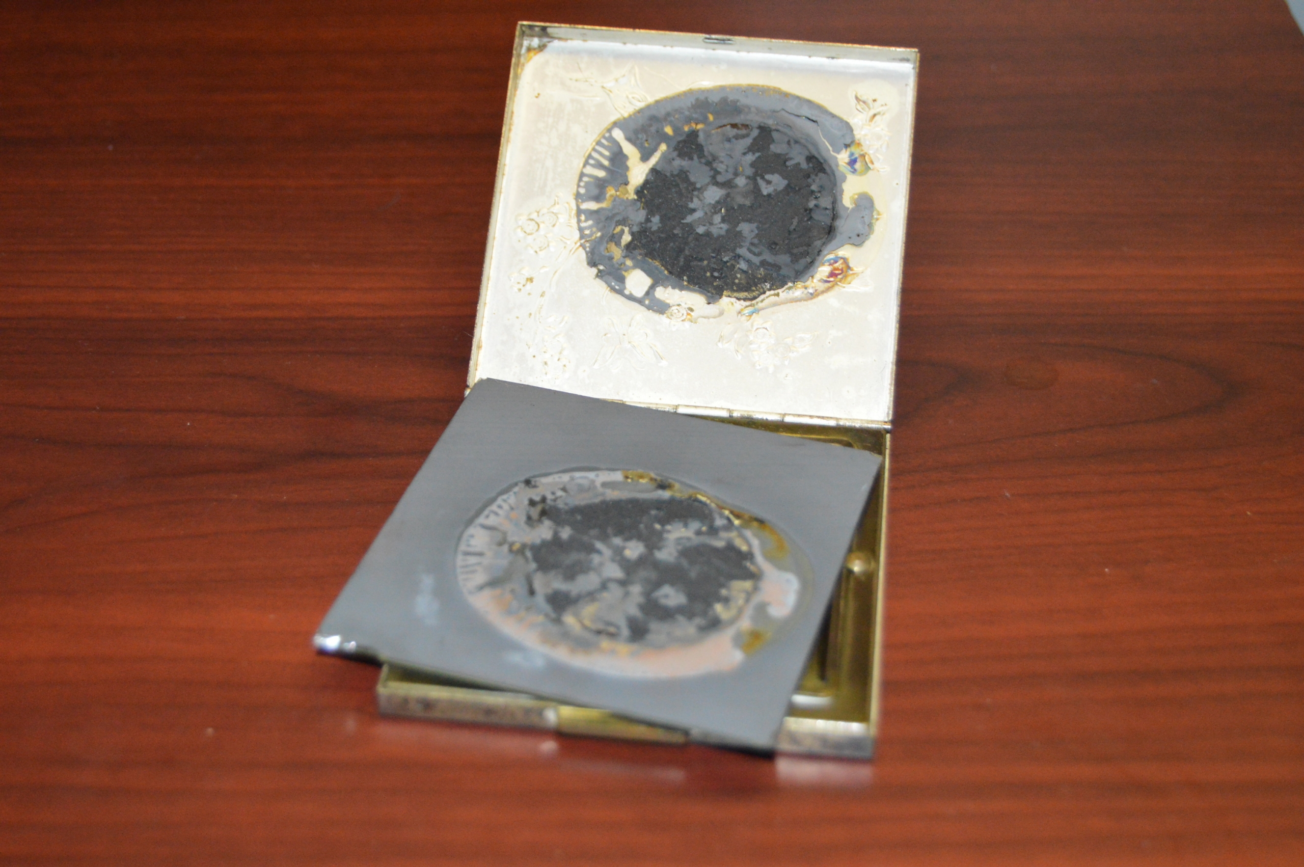 Photo of the small square compact (from Figure 3) with the mirror removed from the top side. Brown and gray residue remains where the mirror was attached.