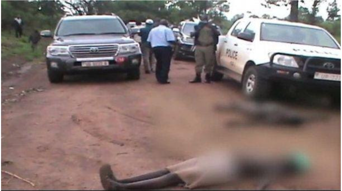 The image depicts women lying down on a dirt road in front of a line of vehicles. The truck on the left of the image is clearly marked "police." The two bodies are blurred to obscure their partially nude bodies