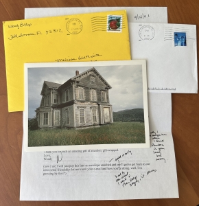 a stack of correspondence: at the top, a card with a photo of an austere, dingy gray against a cloudy gray sky, with a bright yellow addressed envelope underneath. Also visible is another white envelope and, at the bottom of the stack, a typed letter with handwritten additions.